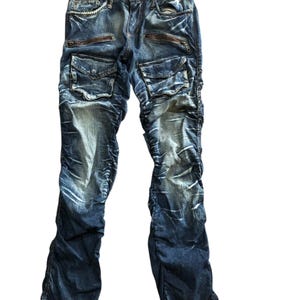 Könnte beinhalten: Blaue Jeans mit mehreren Taschen, Reißverschlüssen und einem Used-Look. Die Jeans haben ein einzigartiges Design mit gerafften Stoffdetails und einem Kettenakzent auf der Vordertasche.