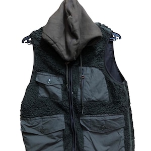 May include: A dark green vest with a hooded collar. The vest has a zip-up front, multiple pockets, and a fuzzy texture. The hood and pockets are a darker shade of green. The vest is hanging on a black hanger.