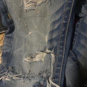 May include: Close-up of distressed blue denim jeans with multiple tears and frayed edges. Silver safety pins are used to repair the rips. The jeans have a faded, worn appearance.