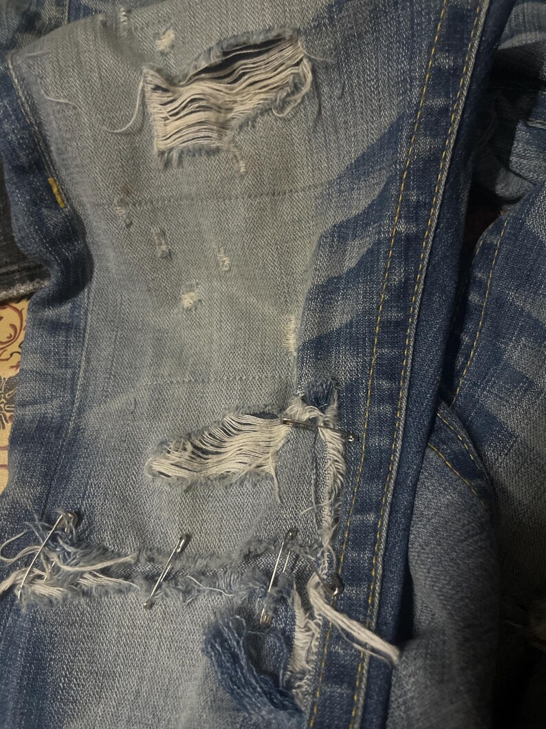 May include: Close-up of distressed blue denim jeans with multiple tears and frayed edges. Silver safety pins are used to repair the rips. The jeans have a faded, worn appearance.