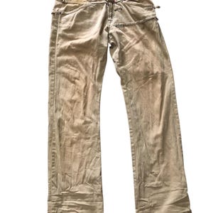May include: Tan denim jeans with a distinctive design. The jeans have a button closure and a unique pocket design. The fabric has a slightly worn appearance. The jeans have a straight leg cut.