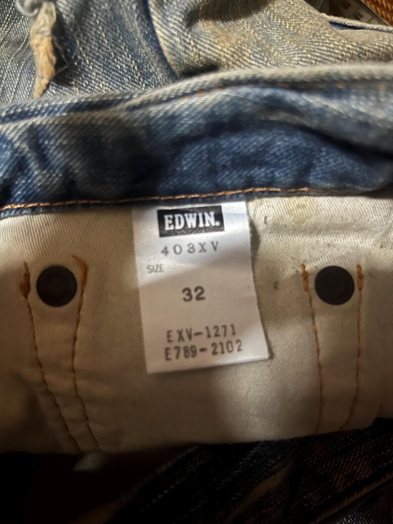 K&ouml;nnte beinhalten: Nahaufnahme der Innenseite einer blauen Jeans. Das Etikett lautet "EDWIN" und enth&auml;lt die Nummern "403XV", "32", "EXV-1271" und "E789-2102". Die Jeans weist Gebrauchsspuren auf.