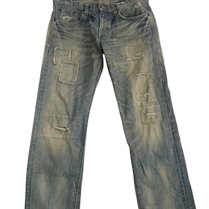 K&ouml;nnte beinhalten: Verblasste blaue Jeans aus Denim mit abgenutzten Details, einschlie&szlig;lich Flicken und Rissen. Die Jeans haben ein klassisches F&uuml;nf-Taschen-Design und einen Knopfverschluss. Das Edwin-Markenetikett ist oben sichtbar.