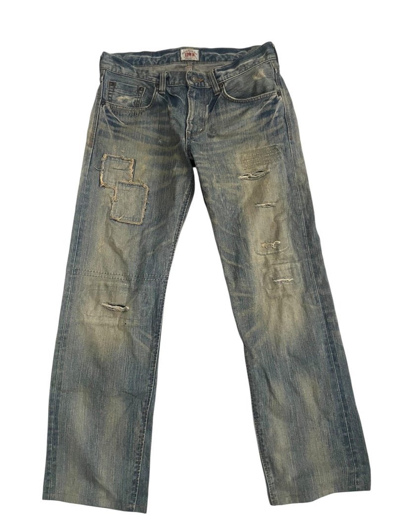 K&ouml;nnte beinhalten: Verblasste blaue Jeans aus Denim mit abgenutzten Details, einschlie&szlig;lich Flicken und Rissen. Die Jeans haben ein klassisches F&uuml;nf-Taschen-Design und einen Knopfverschluss. Das Edwin-Markenetikett ist oben sichtbar.