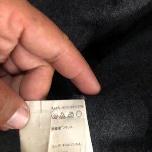 May include: A close-up of a tag with text in Japanese and French, held by a hand. The tag indicates the material composition of the item, with 63% wool and 37% polyester. The tag also includes care symbols and the text "Made in France."
