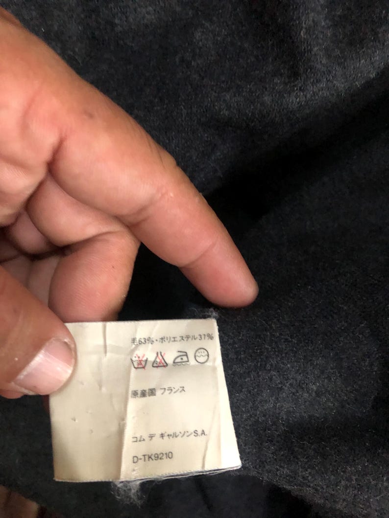 May include: A close-up of a tag with text in Japanese and French, held by a hand. The tag indicates the material composition of the item, with 63% wool and 37% polyester. The tag also includes care symbols and the text "Made in France."