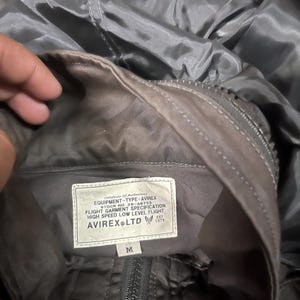 Pu&ograve; includere: Primo piano dell'interno di una giacca grigia con etichetta. L'etichetta recita "EQUIPMENT-TYPE-AVIREX" e "FLIGHT GARMENT SPECIFICATION". La giacca ha una cerniera e la taglia &egrave; "M". La giacca &egrave; realizzata in un materiale lucido e liscio.