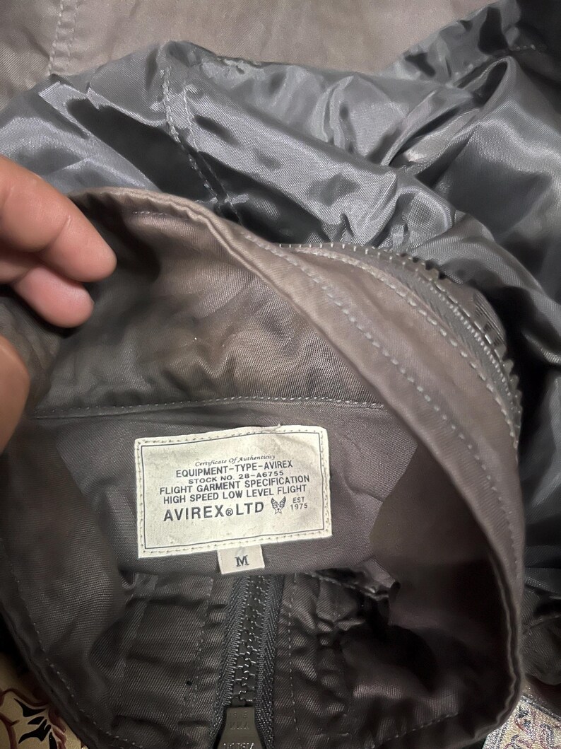 Pu&ograve; includere: Primo piano dell'interno di una giacca grigia con etichetta. L'etichetta recita "EQUIPMENT-TYPE-AVIREX" e "FLIGHT GARMENT SPECIFICATION". La giacca ha una cerniera e la taglia &egrave; "M". La giacca &egrave; realizzata in un materiale lucido e liscio.