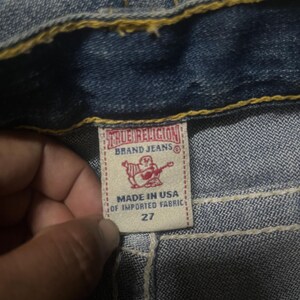 May include: Close-up of a light blue denim garment. The label reads "TRUE RELIGION BRAND JEANS" with a graphic of a Buddha playing a guitar. The label also states "MADE IN USA OF IMPORTED FABRIC" and size 27.