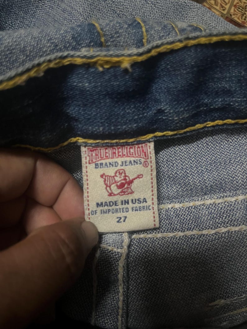 May include: Close-up of a light blue denim garment. The label reads "TRUE RELIGION BRAND JEANS" with a graphic of a Buddha playing a guitar. The label also states "MADE IN USA OF IMPORTED FABRIC" and size 27.