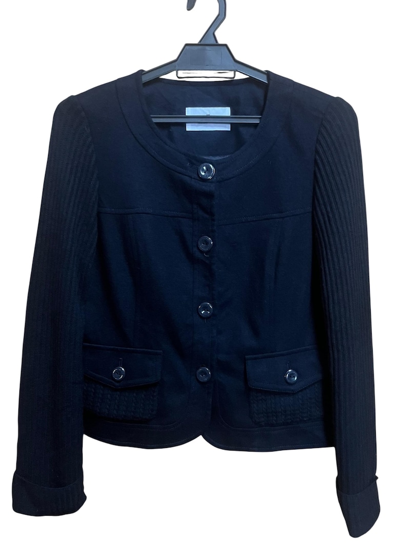 May include: A black jacket with a round neckline and button closure. The jacket has two front pockets and ribbed sleeves. It is hanging on a black hanger.
