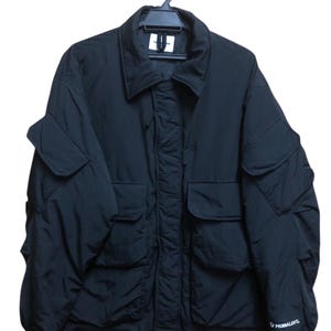 Peut inclure: Veste noire avec plusieurs poches et un col, suspendue à un cintre noir. La veste a un cordon de serrage en bas et le mot "PRIMALOFT" est visible sur la manche. La veste semble être faite d'un matériau léger.