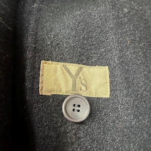 May include: Close-up of a black wool coat. The coat features a small, round, gray button with four holes. A tan label with the text "Y's" is sewn onto the coat. The coat's fabric has a textured appearance.
