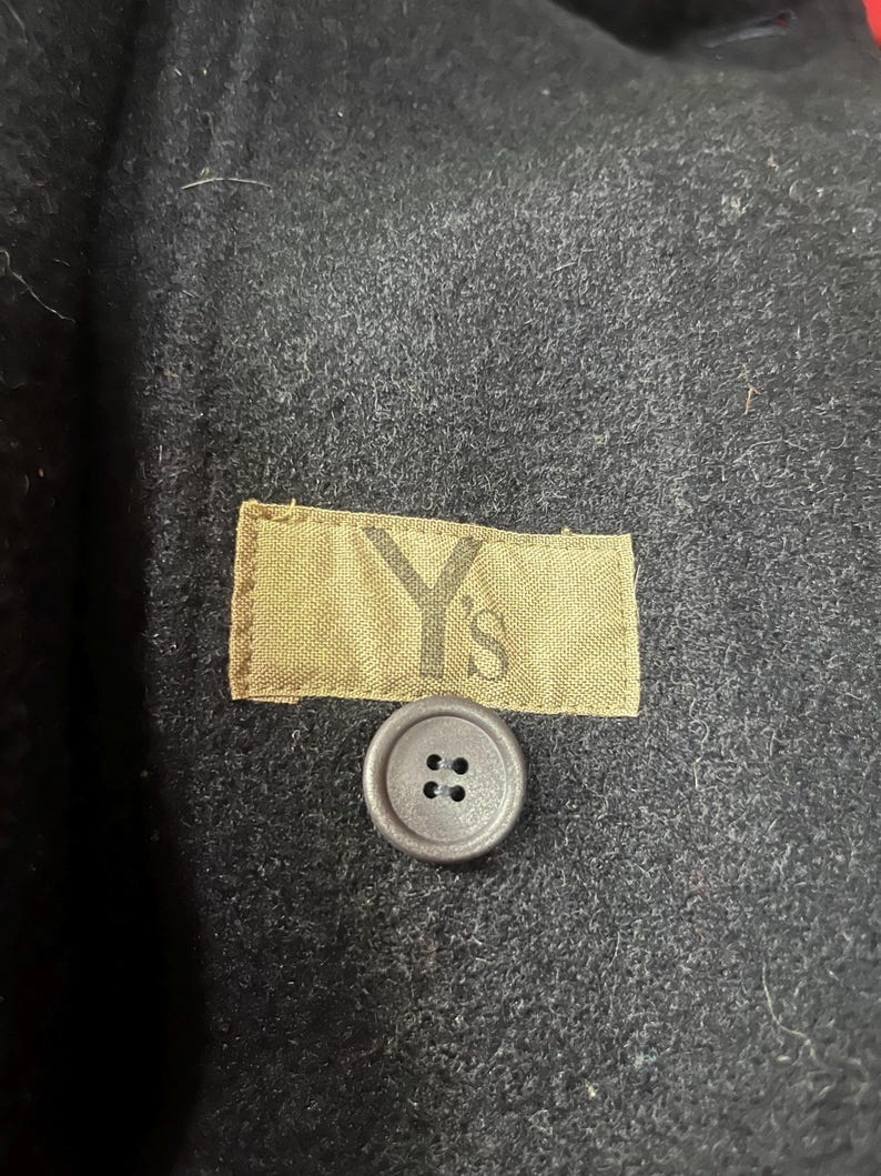 May include: Close-up of a black wool coat. The coat features a small, round, gray button with four holes. A tan label with the text "Y's" is sewn onto the coat. The coat's fabric has a textured appearance.