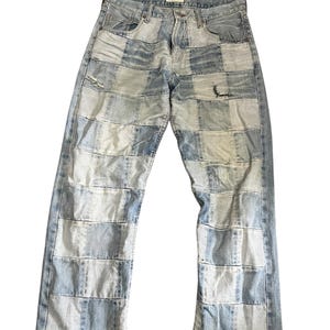 Puede incluir: Pantalones vaqueros de mezclilla azul claro con un diseño de parches compuesto por varios cuadrados rectangulares de tela de mezclilla en tonos alternos de azul claro y medio. Los pantalones incluyen una cintura estándar con trabillas, cierre de botones y detalles desgastados en la zona del muslo.