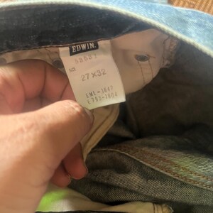 May include: Light blue denim jeans with a visible tag. The tag reads "EDWIN" with the size "27 x 32". The jeans are slightly faded, showing signs of wear. The overall style is casual and classic.