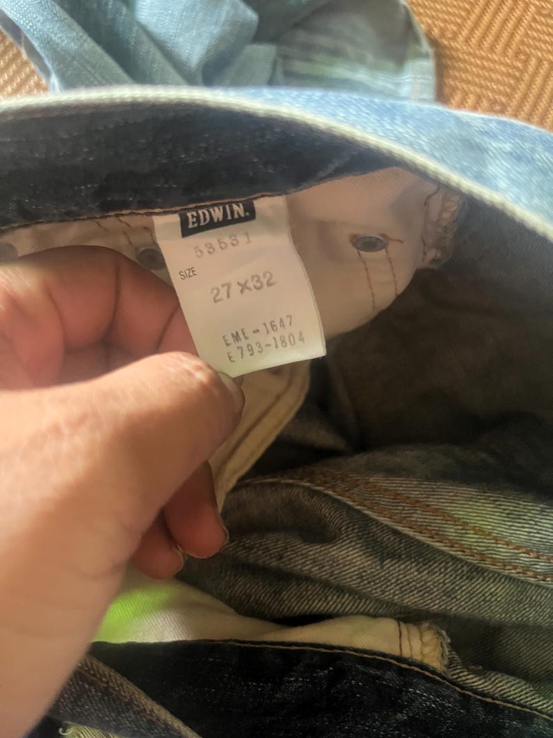 May include: Light blue denim jeans with a visible tag. The tag reads "EDWIN" with the size "27 x 32". The jeans are slightly faded, showing signs of wear. The overall style is casual and classic.