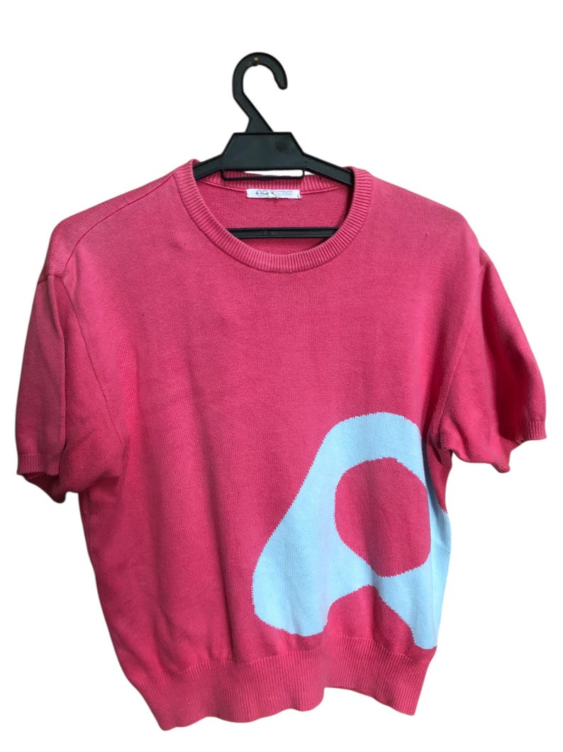 May include: A coral-colored short-sleeved sweater with a large, light blue abstract design on the front. The sweater is hanging on a black hanger against a white background. The design is a large, stylized letter "A".