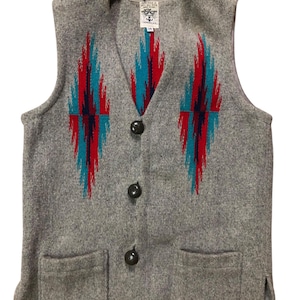 May include: A grey wool waistcoat with a V-neck and two front pockets. The waistcoat features a geometric pattern in red and turquoise. It has four black buttons down the front and a small tag near the neckline.