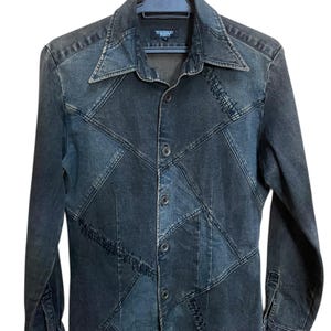 May include: A dark denim button-up shirt with a patchwork design. The shirt features a pointed collar, long sleeves, and a button-down front. The denim is a mix of dark and faded blue tones, creating a geometric pattern. The shirt is hanging on a black hanger.