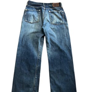 May include: Back view of blue denim jeans with a faded wash. The jeans have two back pockets and a brown leather patch at the waistband. The denim is a medium wash with some lighter areas.