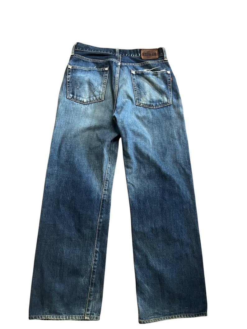 May include: Back view of blue denim jeans with a faded wash. The jeans have two back pockets and a brown leather patch at the waistband. The denim is a medium wash with some lighter areas.