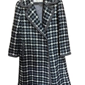 May include: A long, houndstooth-patterned coat in black, white, and gray, with a gray collar and trim. The coat has a classic design and is hanging on a black hanger. The label reads "MISS BALMAIN".