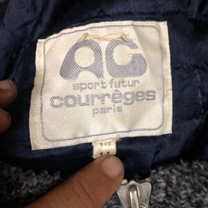 May include: Close-up of a navy blue jacket label with the text "AC sport futur courr&egrave;ges paris". The label is white with a light blue logo. A silver zipper is visible at the bottom.