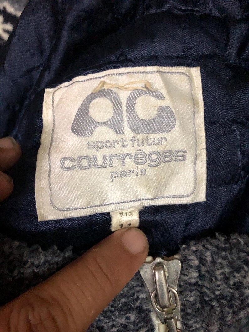 May include: Close-up of a navy blue jacket label with the text "AC sport futur courr&egrave;ges paris". The label is white with a light blue logo. A silver zipper is visible at the bottom.