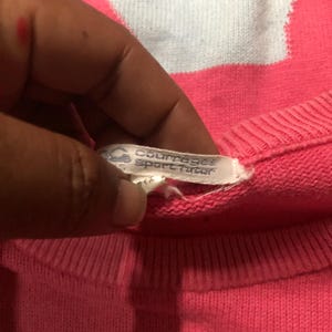 May include: Close-up of a pink Courreges Sport Futur sweater. The tag is visible, showing the brand name. The sweater is a solid pink color with a ribbed neckline.