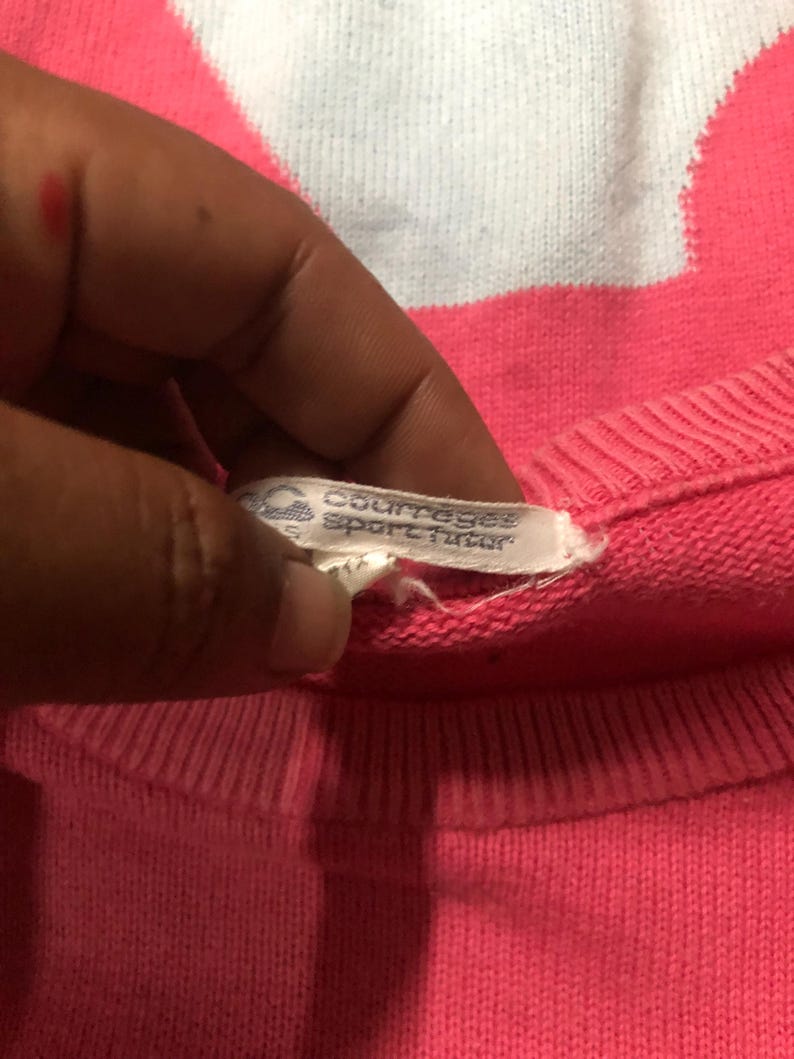 May include: Close-up of a pink Courreges Sport Futur sweater. The tag is visible, showing the brand name. The sweater is a solid pink color with a ribbed neckline.