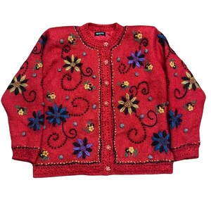 May include: A bright red long-sleeve knit cardigan featuring a crew neckline and a front button closure. The garment is decorated with embroidered floral patterns in blue, yellow, and black, accented by small beads and swirling black thread details.