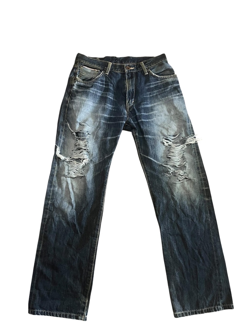 May include: Dark wash denim jeans with distressed details. The jeans have multiple rips and tears across the front of the legs. The denim has a faded, worn appearance.