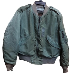 Könnte beinhalten: Eine grüne Bomberjacke mit Reißverschluss, zwei Vordertaschen und einer kleinen Tasche am linken Ärmel. Die Jacke hat einen gerippten Kragen, Bündchen und Saum. Sie hängt an einem schwarzen Kleiderbügel vor weißem Hintergrund.