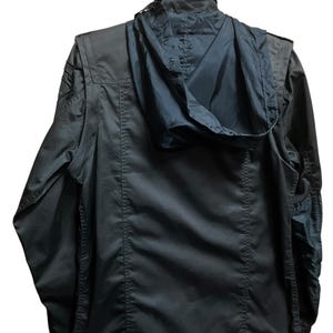 Pu&ograve; includere: Giacca verde oliva scuro con cappuccio nero, appesa a una gruccia nera. La giacca ha una chiusura a cerniera, tasche multiple e una toppa in stile militare sulla spalla. Il cappuccio &egrave; ripiegato. La giacca &egrave; realizzata in un materiale resistente e idrorepellente.