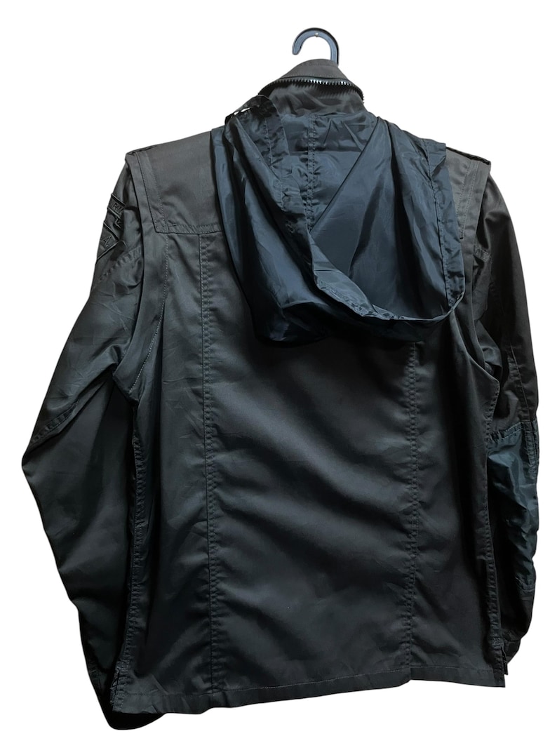 Pu&ograve; includere: Giacca verde oliva scuro con cappuccio nero, appesa a una gruccia nera. La giacca ha una chiusura a cerniera, tasche multiple e una toppa in stile militare sulla spalla. Il cappuccio &egrave; ripiegato. La giacca &egrave; realizzata in un materiale resistente e idrorepellente.