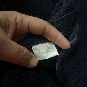 May include: Close-up of a dark navy blue fabric with a white tag. The tag reads "MADE IN JAPAN" and lists the fabric composition as 90% wool and 10% nylon. The fabric appears to be part of a garment.