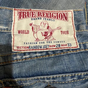 May include: Blue denim jeans featuring a white rectangular patch with red text and a graphic of a seated figure playing a guitar. The patch reads True Religion Brand Jeans, World Tour, Fashion for the Senses, Section Rainbow Joey Row 28 Seat 33.