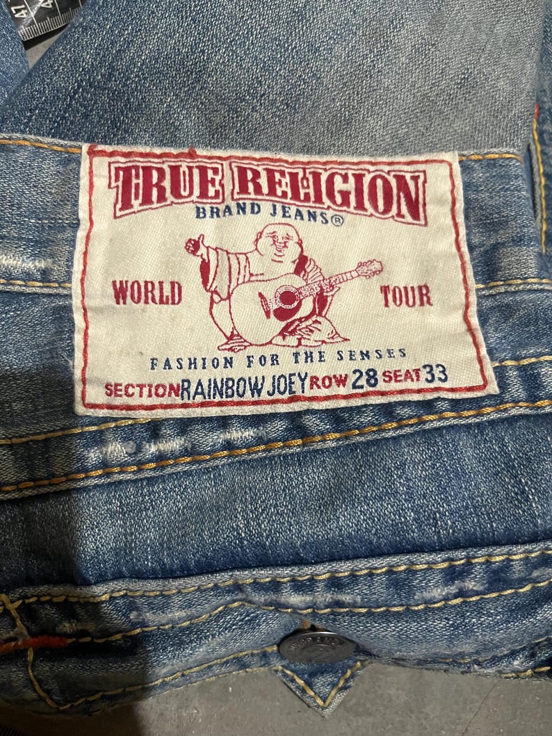 May include: Blue denim jeans featuring a white rectangular patch with red text and a graphic of a seated figure playing a guitar. The patch reads True Religion Brand Jeans, World Tour, Fashion for the Senses, Section Rainbow Joey Row 28 Seat 33.