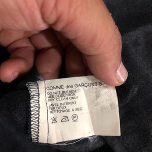 May include: A close-up of a tag from a Comme des Gar&ccedil;ons garment. The tag is white with black text, including care instructions such as "DO NOT BLEACH" and symbols for ironing and dry cleaning. The tag is attached to a dark gray fabric.