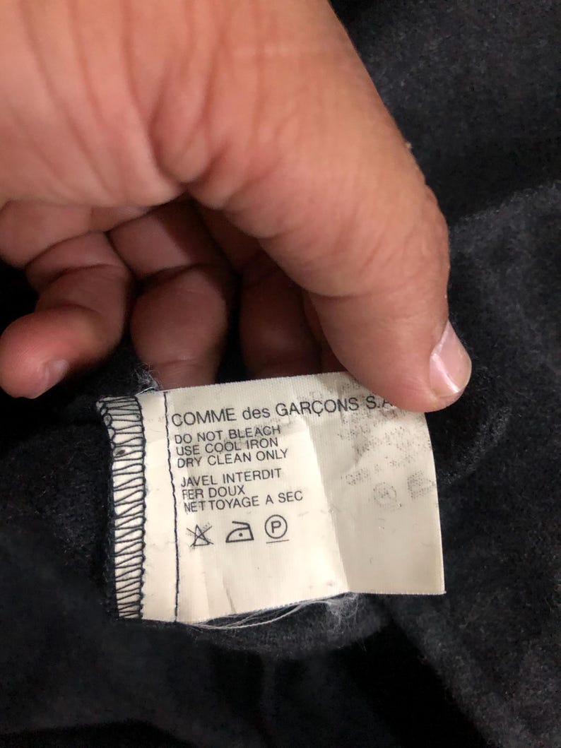 May include: A close-up of a tag from a Comme des Gar&ccedil;ons garment. The tag is white with black text, including care instructions such as "DO NOT BLEACH" and symbols for ironing and dry cleaning. The tag is attached to a dark gray fabric.