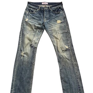 May include: Blue denim jeans featuring a distressed finish with frayed areas, whiskering patterns, and visible stitched patches on the front legs. The jeans include a button fly closure, five pockets, and a pink interior label at the waistband.