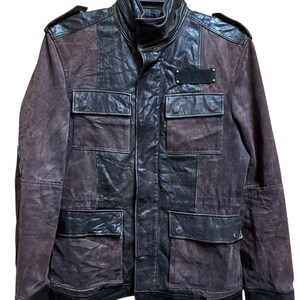 May include: A brown and black leather jacket with a high collar and multiple pockets. The jacket features a mix of textures, with smooth and distressed leather panels. The design includes shoulder epaulettes and a small, dark patch on the chest.