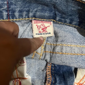 May include: Blue denim jeans featuring thick orange contrast stitching and a white interior label with the red True Religion logo and the text made in USA. The inner pocket lining displays a repeating red graphic print of the brand logo.