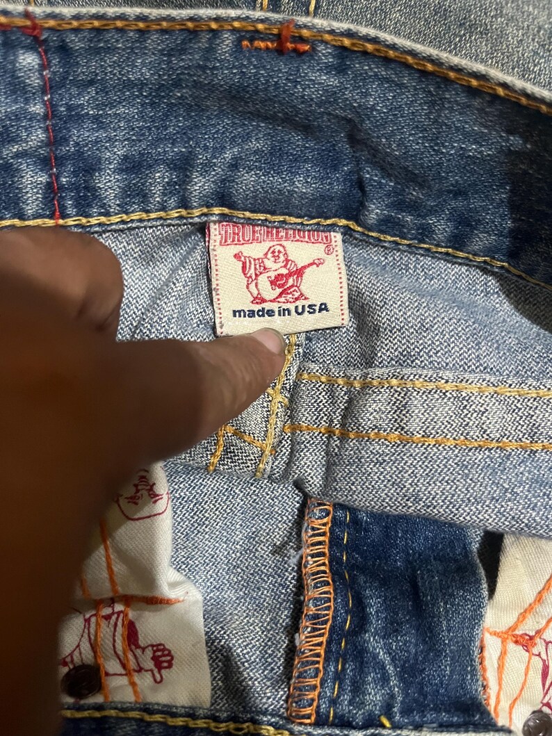 May include: Blue denim jeans featuring thick orange contrast stitching and a white interior label with the red True Religion logo and the text made in USA. The inner pocket lining displays a repeating red graphic print of the brand logo.