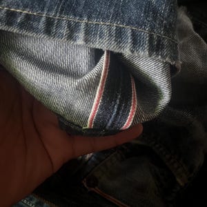 May include: Close-up of denim jeans, showcasing the inside seam with a red and white stripe detail. The jeans are a medium wash blue with visible stitching and a worn appearance, suggesting a vintage or well-loved item.
