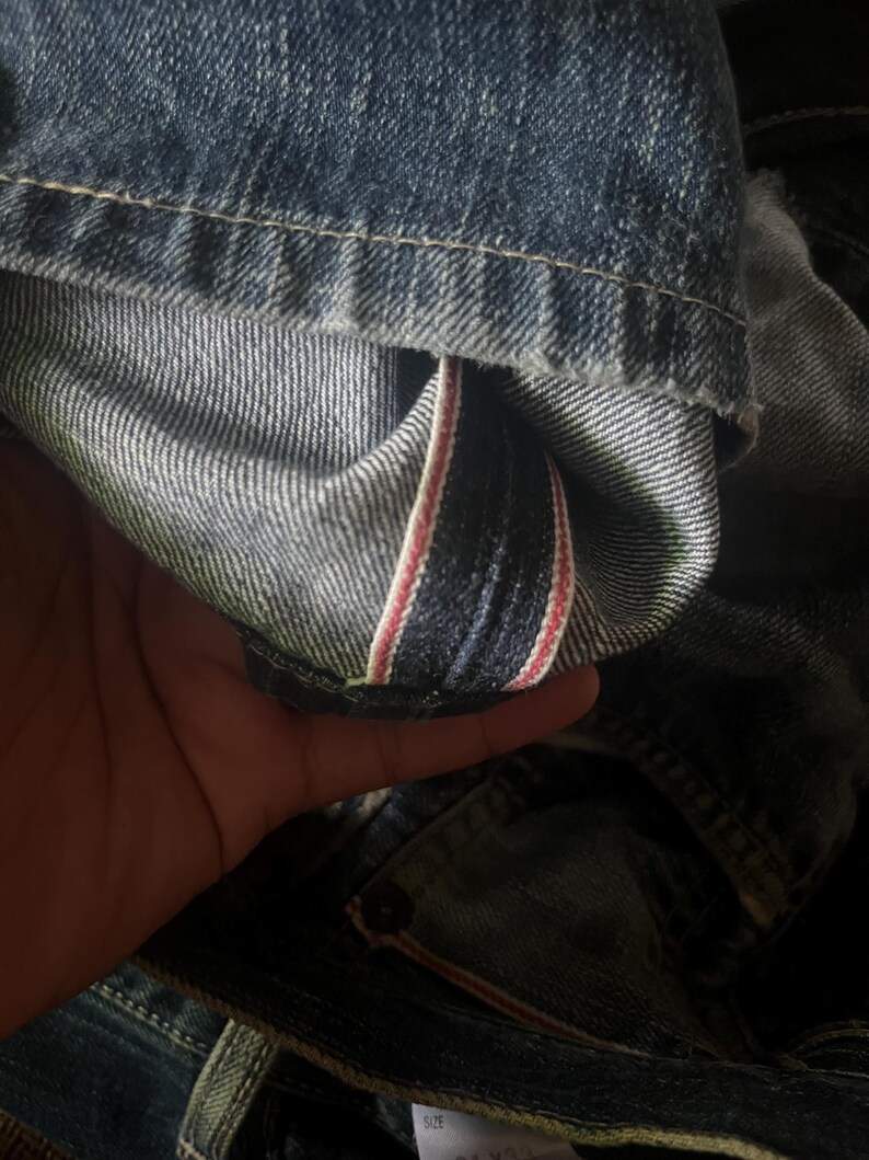 May include: Close-up of denim jeans, showcasing the inside seam with a red and white stripe detail. The jeans are a medium wash blue with visible stitching and a worn appearance, suggesting a vintage or well-loved item.