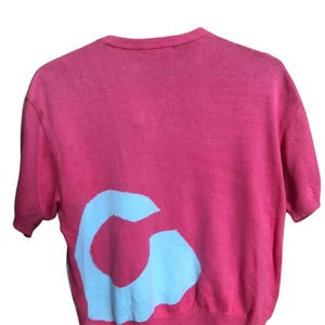 May include: A short-sleeved, coral-colored sweater with a white graphic on the back. The sweater is hanging on a black hanger. The graphic is a stylized letter "G".