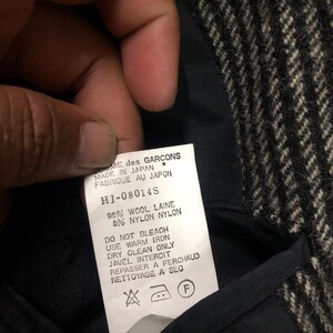 May include: Close-up of a garment label with text. The label reads "COMME des GARCONS MADE IN JAPAN" and includes care instructions. The fabric appears to be a black and white herringbone pattern.