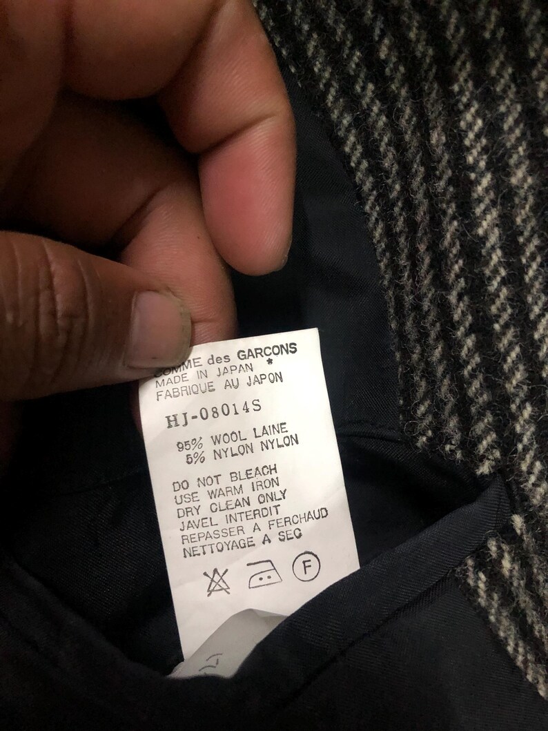 May include: Close-up of a garment label with text. The label reads "COMME des GARCONS MADE IN JAPAN" and includes care instructions. The fabric appears to be a black and white herringbone pattern.